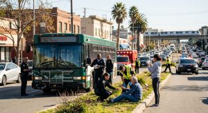 Professional photograph of bus accident aftermath in Oakland, California, showing first responders assisting victims, damaged bus, and emergency vehicles.